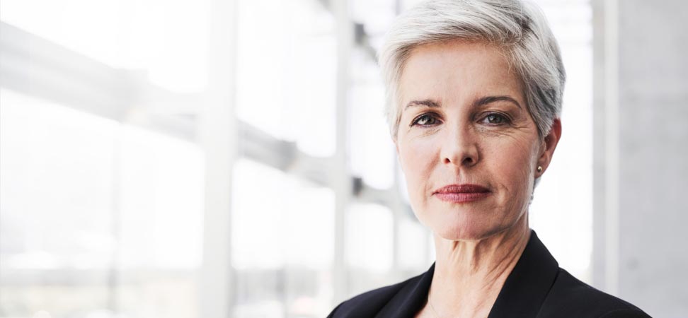 Une femme d’affaires aux cheveux blancs est debout près de fenêtres de bureaux lumineuses.